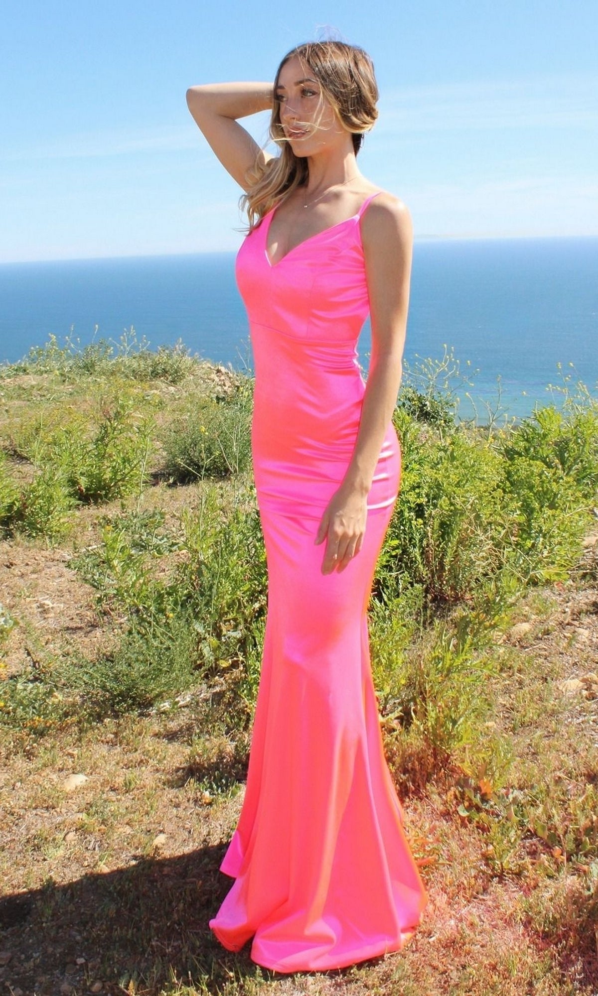 A woman wearing the Backless Neon Tight Long Prom Dress SE016 stands outdoors on grassy terrain, with the ocean and blue sky in the background.